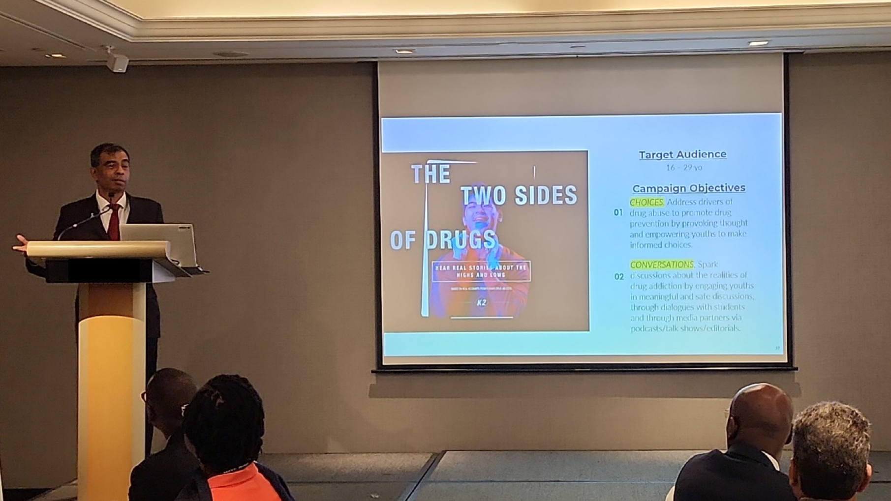 Speaker at podium presenting "The Two Sides of Drugs" campaign objectives on projector screen to audience.